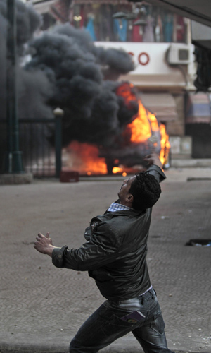 Manifestante joga pedra na polícia nesta sexta-feira (28) no Cairo; ao fundo, um carro policial em chamas