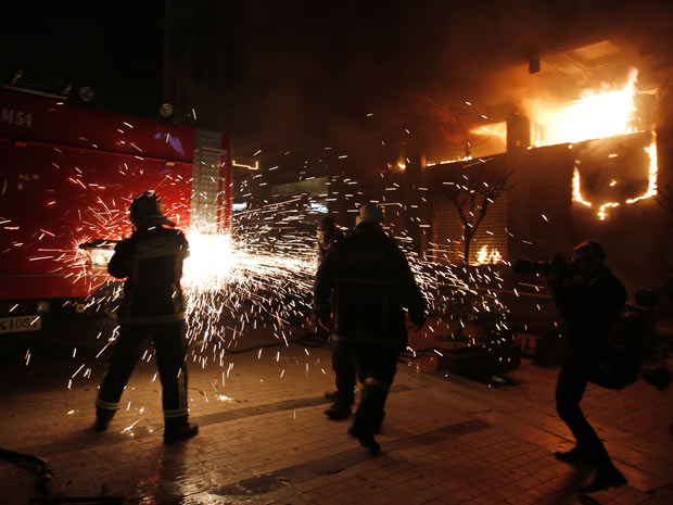 Bombeiros tentam abrir com serra-elétrica caminhão utilizado para conter incêndios, já que manifestantes tomaram as chaves durante protesto realizado em Atenas. À direita, loja é consumida pelo fogo (Foto: Thanassis Stavrakis/AP)