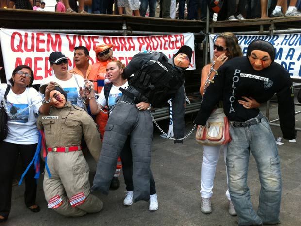 Manifestantes se acorrentam a bonecos vestidos com fardas da polícia e dos bombeiros (Foto: Carolina Lauriano/ G1)